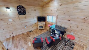 A room with wooden walls, a dartboard, a television, a gaming console, and a red stool.