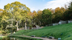 A serene lakeside setting with a wooden cabin, a stone bench, and a grassy slope. - Secluded Woodland Cabin in North Cumbria (Brampton)