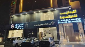 Un edificio con linee gialle e blu illuminate, un cartello che indica Alfursan United Serviced Apartments e diverse auto parcheggiate davanti.