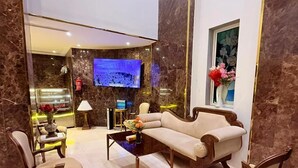 Spaziosa hall con un grande divano, poltrone e un tavolino da caffè. Una televisione è montata a parete, e su un tavolo c'è un vaso con fiori.