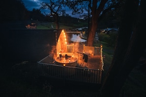 Un espace avec un jacuzzi, doté d’une terrasse en bois et d’un petit bâtiment, le tout entouré d’arbres.