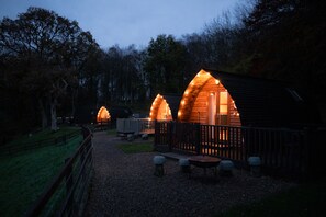 Una hilera de cabañas de madera con iluminación cálida, rodeadas por un camino de grava y una valla de madera.