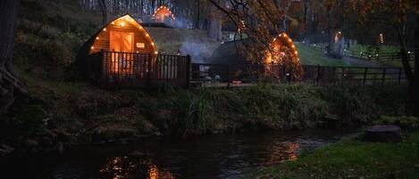 Filera de cabanes de fusta amb finestres il·luminades, un petit rierol i un fons d'arbres.