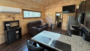 A cozy cabin interior with a wooden ceiling, a sofa, a coffee station, and a kitchen area.