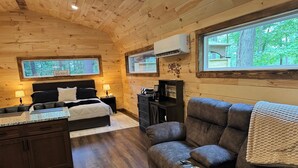 A cozy cabin interior with a bed, a sofa, a small kitchen area, and a window with a view of trees.