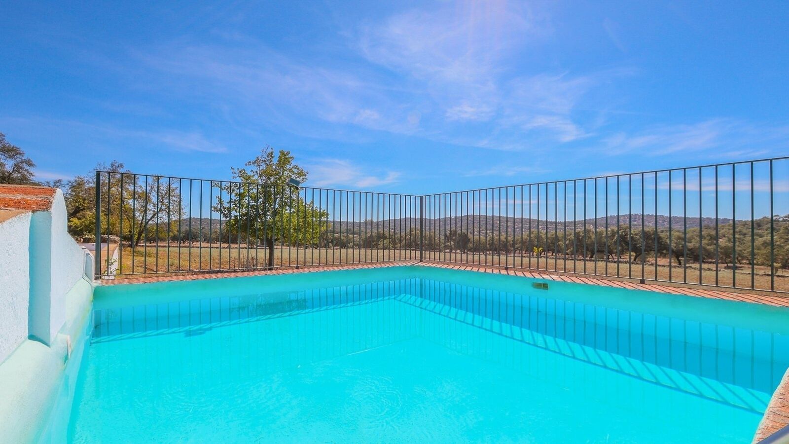 Piscina rectangular con agua cristalina azul, rodeada por una cerca de metal, con árboles y colinas al fondo.