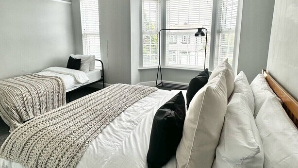 A hotel room with a large bed, a smaller bed, and a desk. There are multiple windows with blinds, a hanging light fixture, and a view of buildings outside. - Luxury 5 bed salford home (free parking) (Salford)