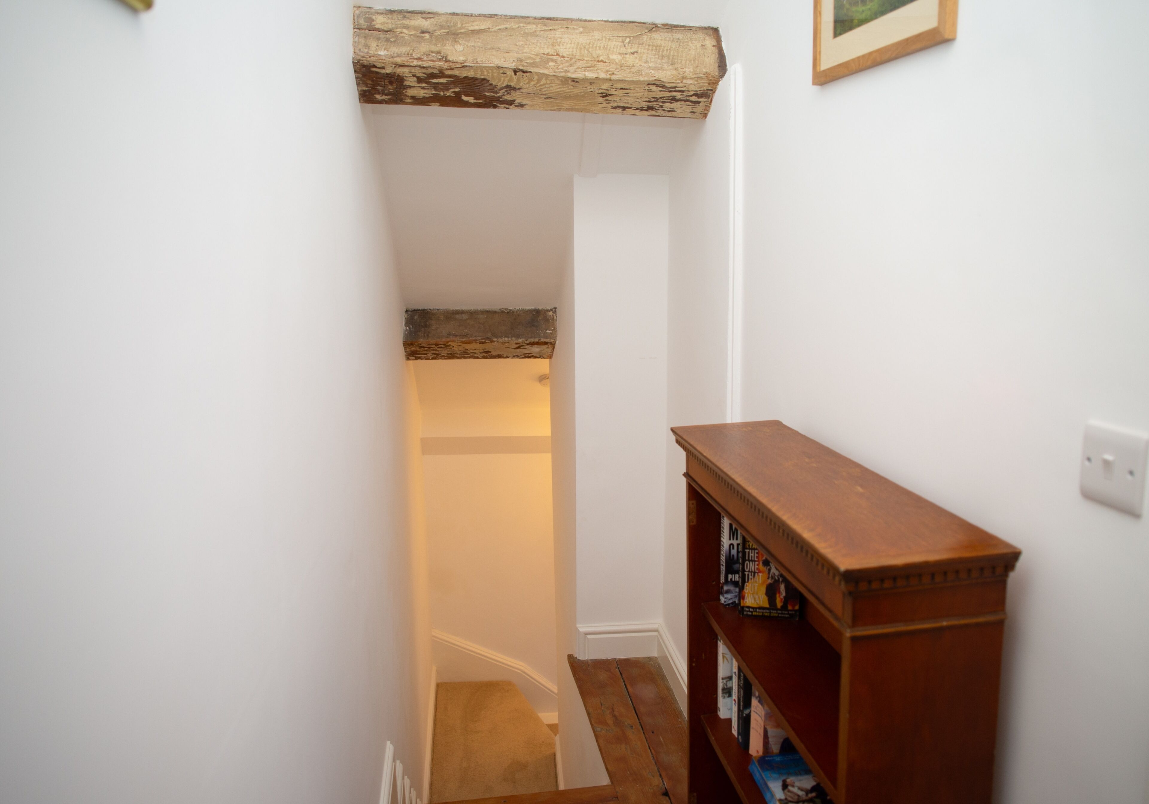 A narrow staircase with a wooden beam ceiling, a wooden bookshelf, and a framed picture on the wall.