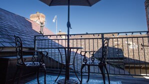 A balcony with a table and two chairs, a large umbrella, and a view of a brick building with a chimney.