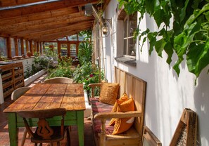 A wooden table and bench set under a wooden roof, surrounded by potted plants and a white wall.