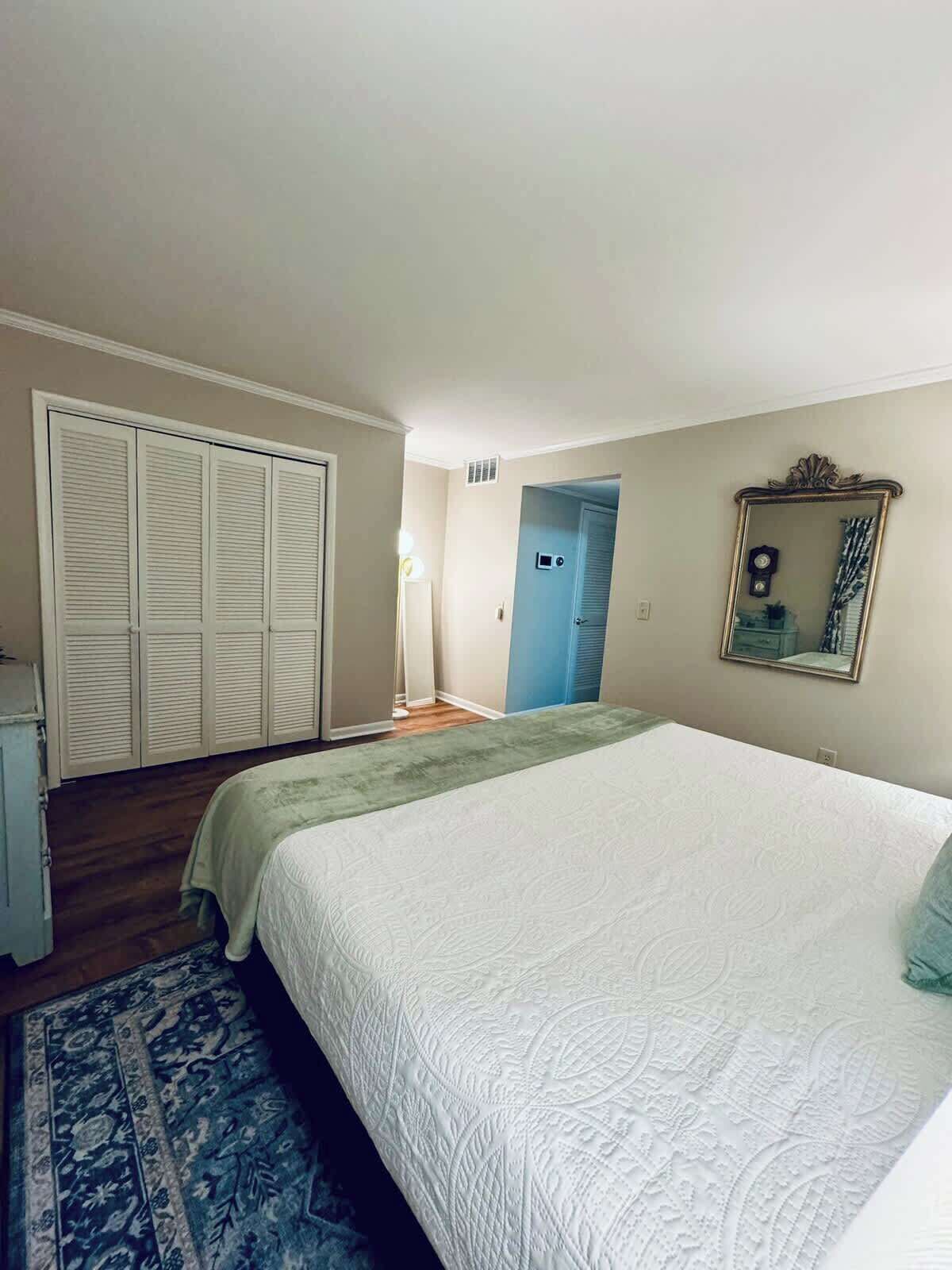 Een slaapkamer met een groot bed, een kledingkast, een spiegel en een deur naar een andere kamer.