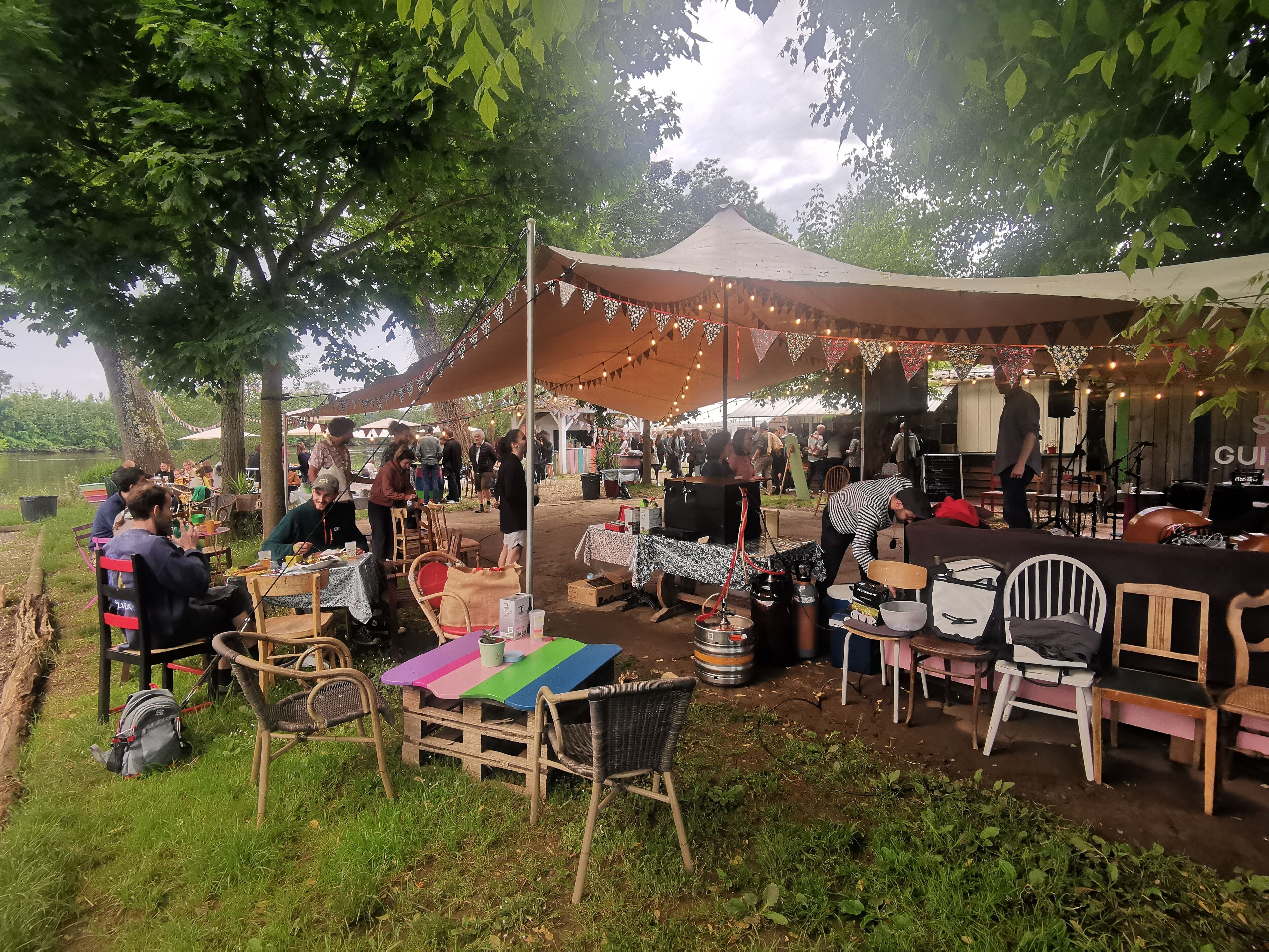 A market stall with tables and chairs, surrounded by trees and a tent.