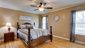 Un dormitorio con cama de madera, una mesita de noche, un ventilador de techo, un reloj y una ventana con cortinas.