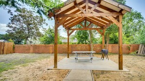 Un pabellón de madera con mesa de picnic y parrilla, rodeado de un patio cercado y árboles.