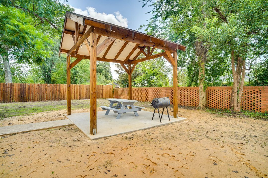 Una pérgola de madera con una mesa de picnic y una parrilla de barbacoa en un patio con suelo arenoso y una cerca de madera.