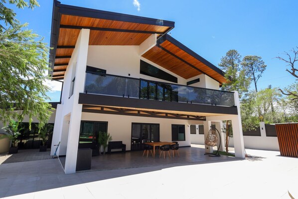 A modern two-story house with a balcony, large windows, and a patio area. - Gey Tropical House (Liberia)