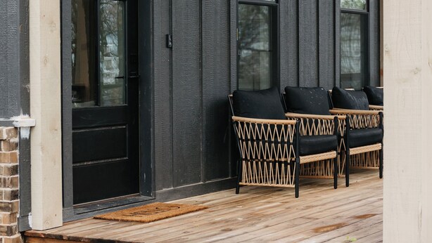 Une véranda en bois avec une porte noire et deux fauteuils en osier dotés de coussins noirs.