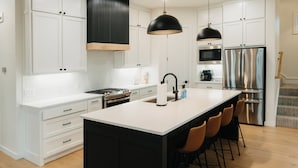 Une cuisine moderne avec des meubles blancs, un îlot central doté de tabourets de bar, des appareils en acier inoxydable et des luminaires suspendus noirs.