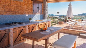Uma cozinha externa com parede de pedra, armários de madeira e uma mesa com tigelas e frutas.