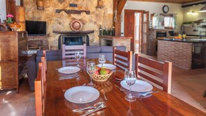 Uma sala de jantar com uma mesa de madeira posta para quatro pessoas, cercada por cadeiras de madeira. Uma parede de pedra e uma lareira são visíveis ao fundo.