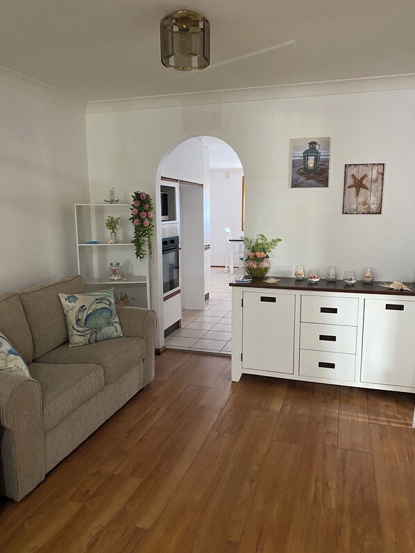 A modern kitchen with white cabinets, a wooden countertop, and a built-in oven. There is a white dining area with a chair and a small table. The room has wooden flooring and a ceiling light fixture.