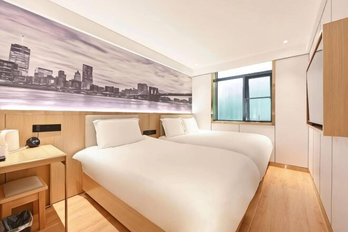 A hotel room with a large bed, a desk with a chair, a small seating area, a window with a view, and a cityscape painting on the wall.