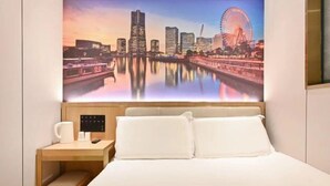 Habitación de hotel con cama, mesita de noche, cafetera y una pintura de paisaje urbano en la pared.