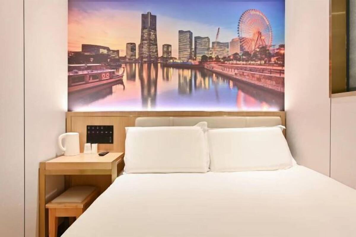 A hotel room with a bed, a nightstand, a coffee maker, and a cityscape painting on the wall.