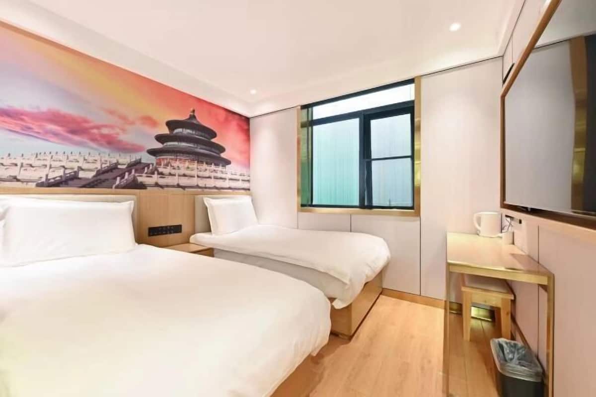 A hotel room with two beds, a desk, a trash bin, a window, and a mural of a traditional building.