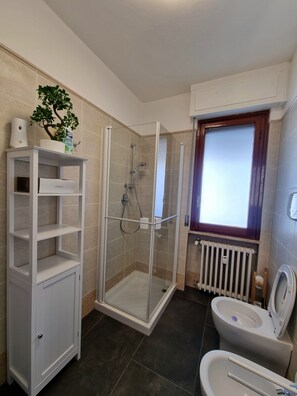 A bathroom with a glass-enclosed shower, a white shelving unit, a toilet, and a radiator.