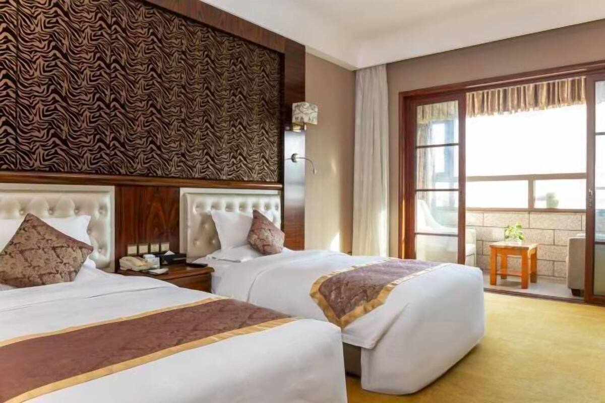 A hotel room with two beds, a patterned wall, a window with curtains, and a small table with a plant.