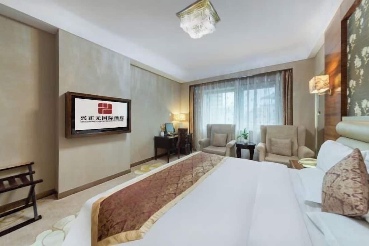 A hotel room with a large bed, a desk, two chairs, a TV mounted on the wall, and a window with curtains.