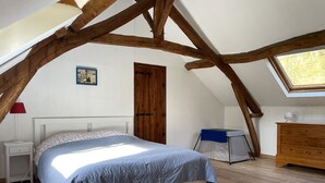 Une chambre avec un plafond en pente, un lit recouvert d’une courtepointe bleue, une commode en bois, une table de chevet avec une lampe et une fenêtre donnant sur les arbres.