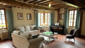 Un salon chaleureux avec un canapé, un fauteuil et une table sur laquelle est posé un jeu de société. On y trouve également un piano et un radiateur.