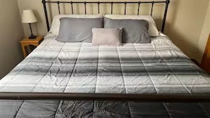 Een bed met een metalen frame, een grijs-wit gestreept dekbed en twee kussens.