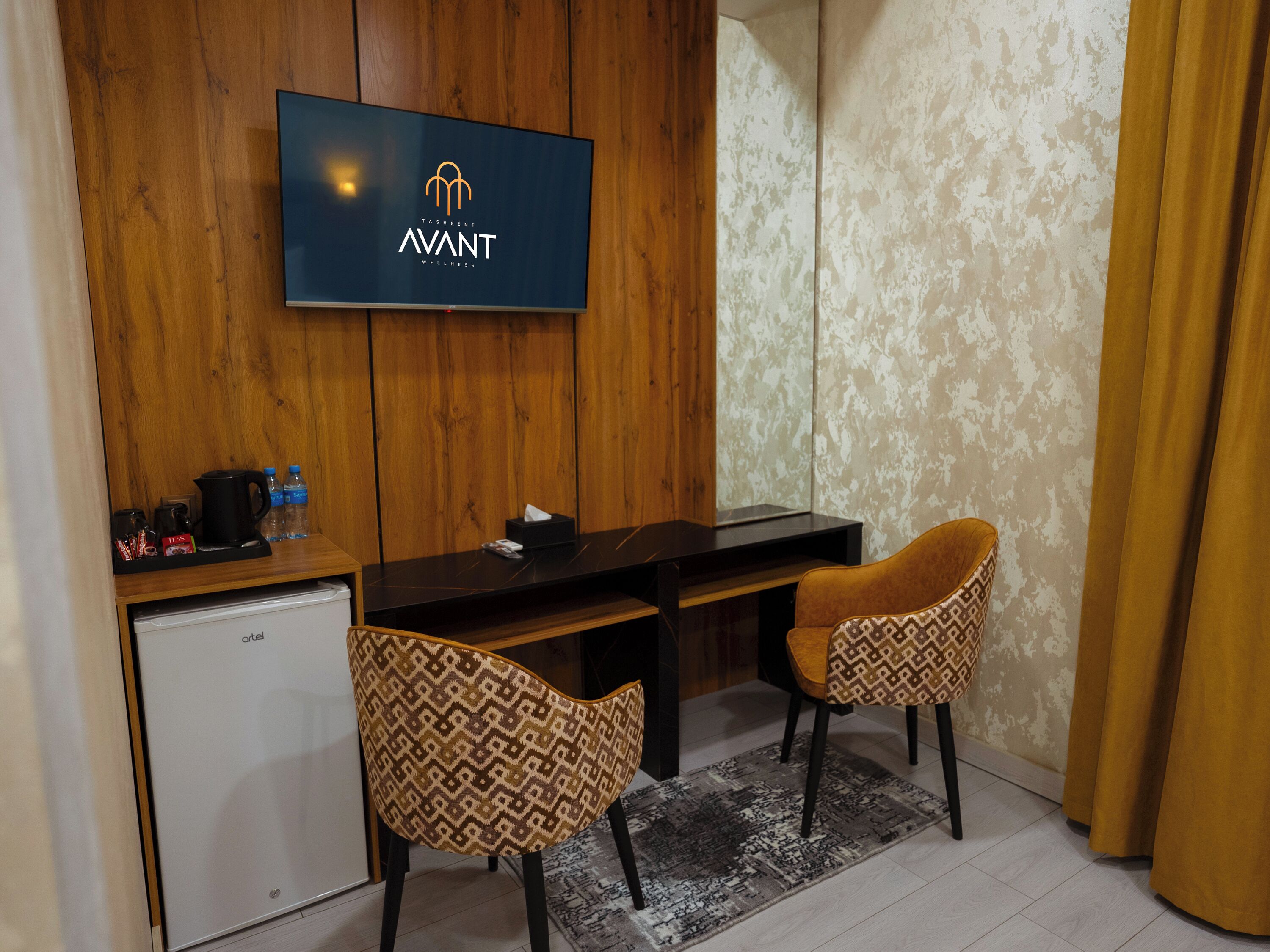 A hotel room with a wooden wall, a flat-screen TV mounted above a desk, two patterned chairs, a small refrigerator, and a patterned rug.