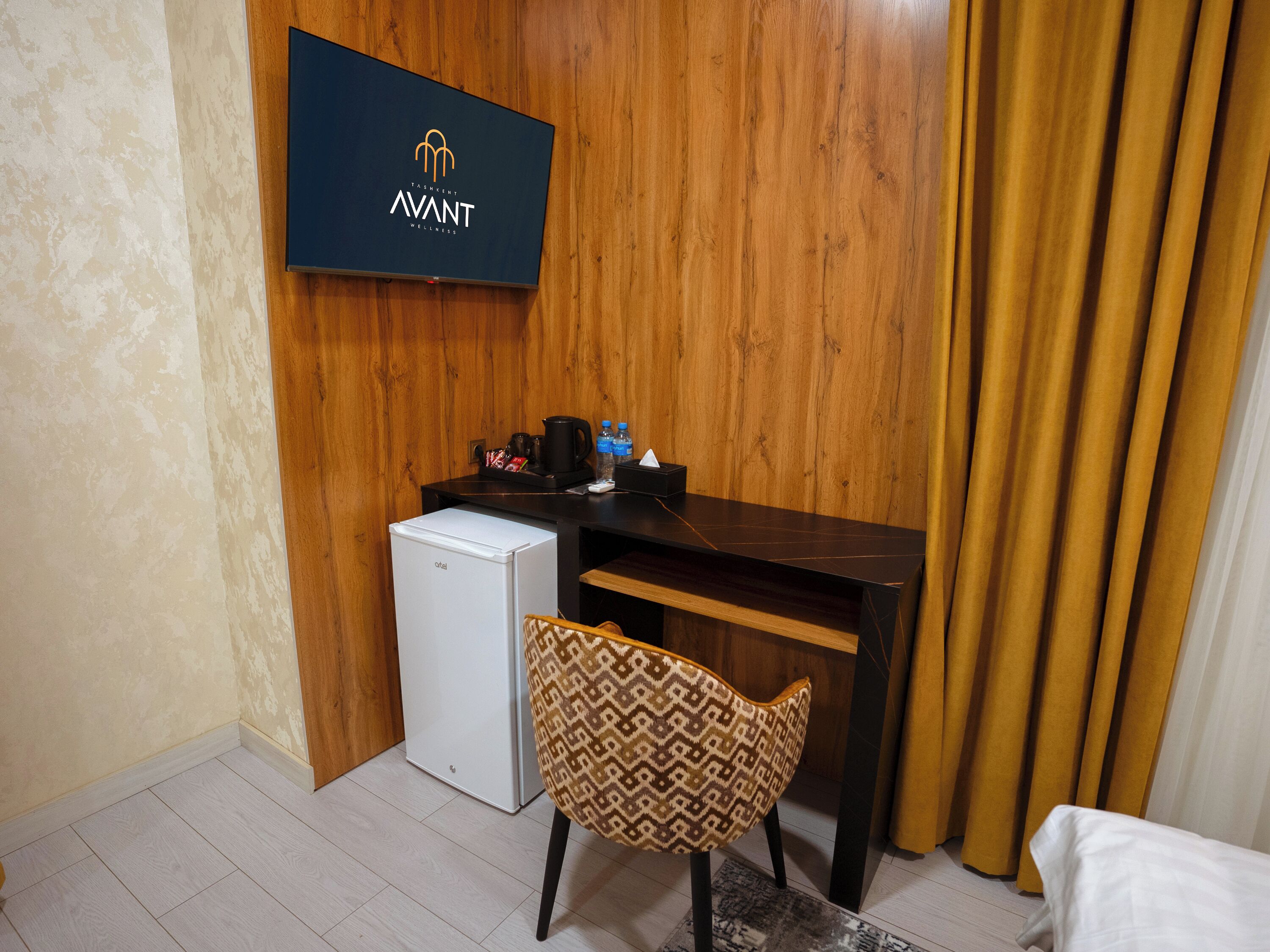 A hotel room with a wooden wall, a television displaying the 'AVANT' logo, a small white refrigerator, a patterned chair, and a bed with a white cover.