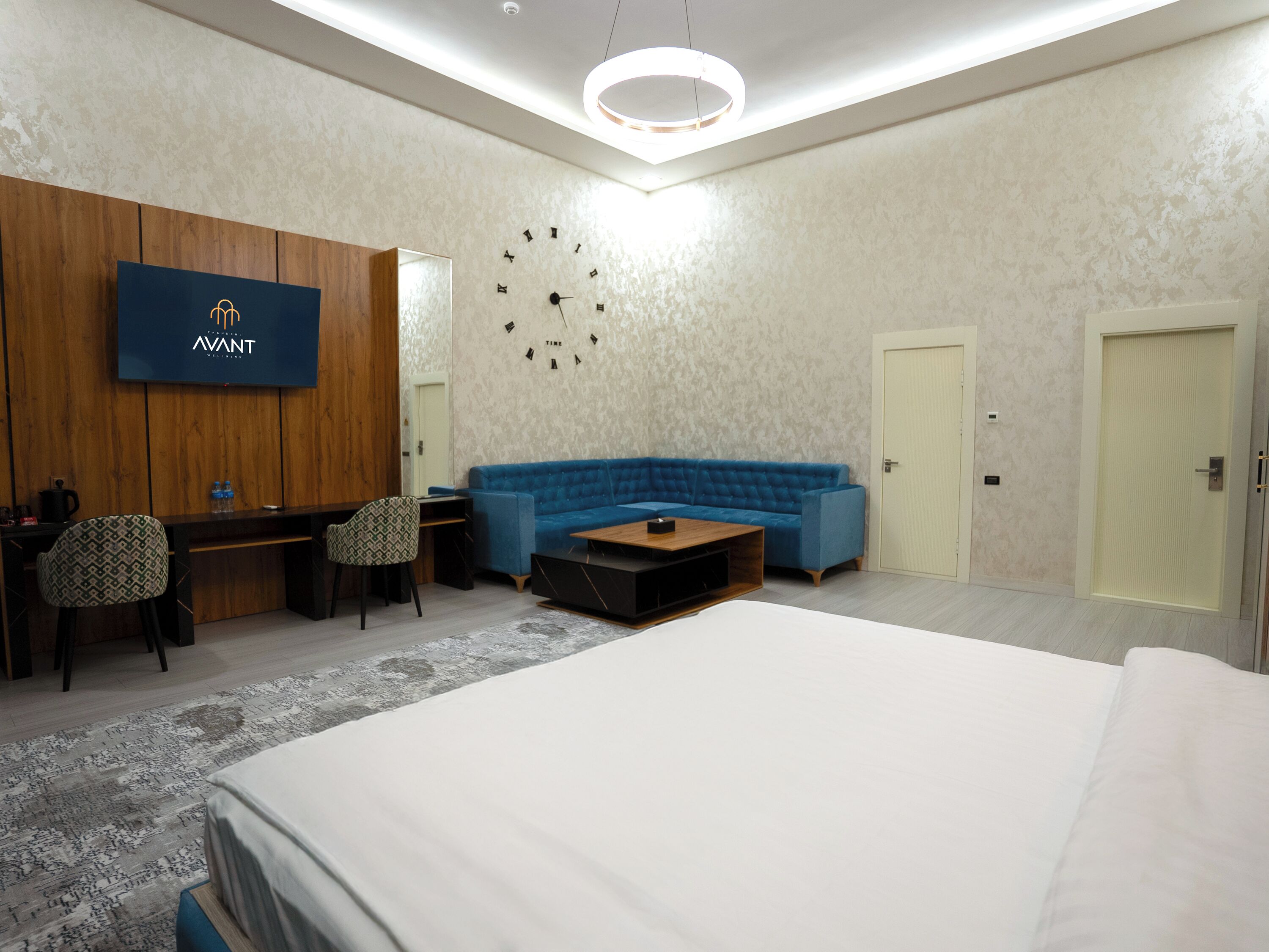 A modern hotel room with a large bed, a blue sofa, a wooden desk, and a TV mounted on the wall.
