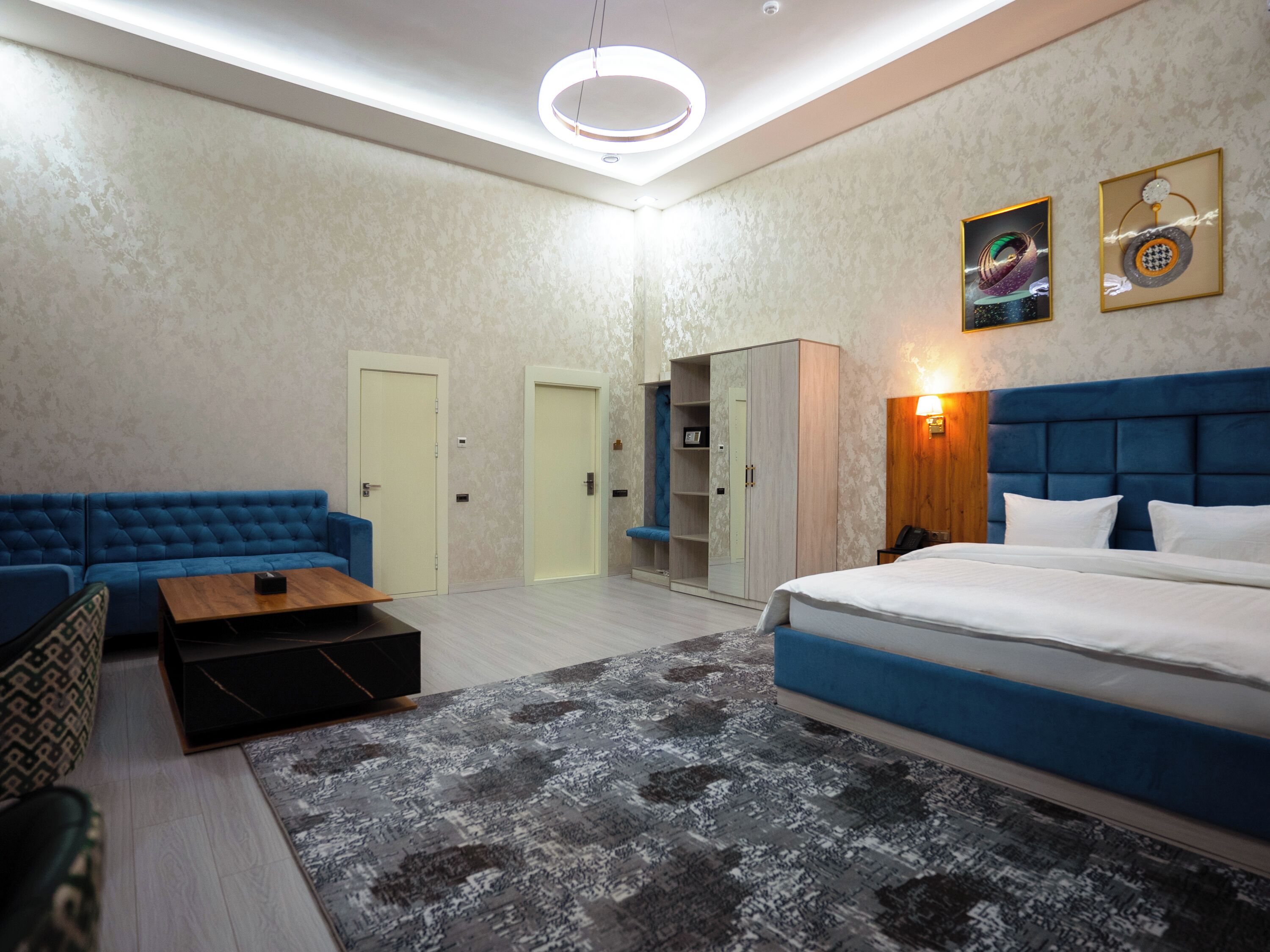 Quarto de hotel moderno com sofá azul, mesa de centro de madeira, cama grande com roupa de cama branca e tapete estampado. Há duas portas, um guarda-roupa e obras de arte penduradas na parede.