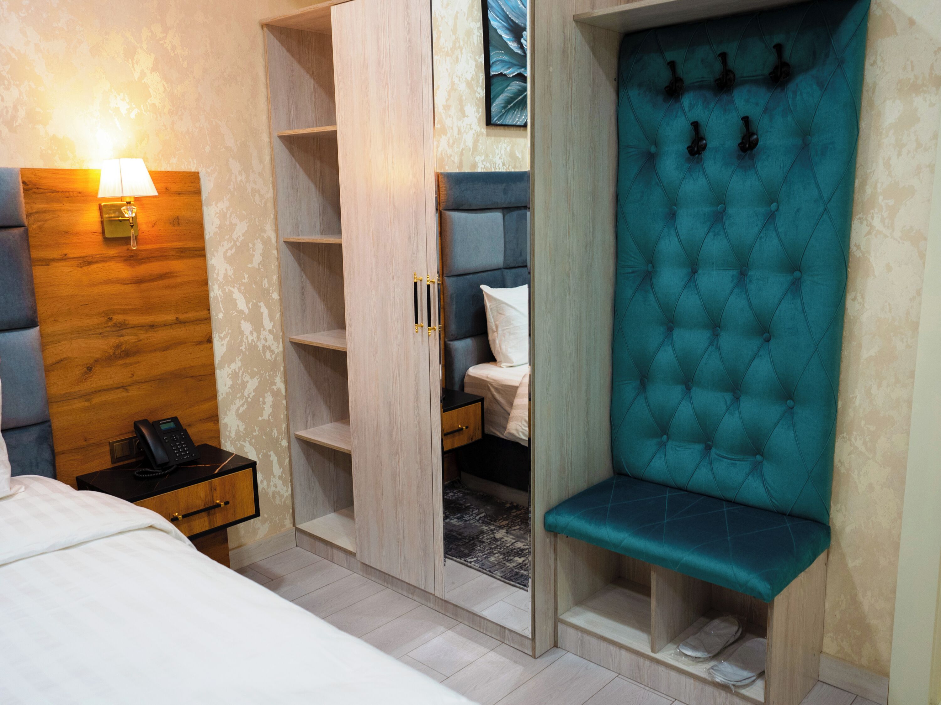 Habitación de hotel con cama, mesita de noche con lámpara, armario con varios estantes, banco con ganchos y cabecera con diseño acolchado.