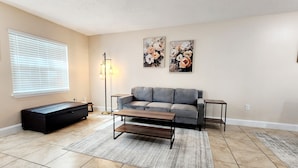 A living room with a grey sofa, a coffee table, a floor lamp, and two floral paintings on the wall. - Toria Suites 922 (Lakeland)