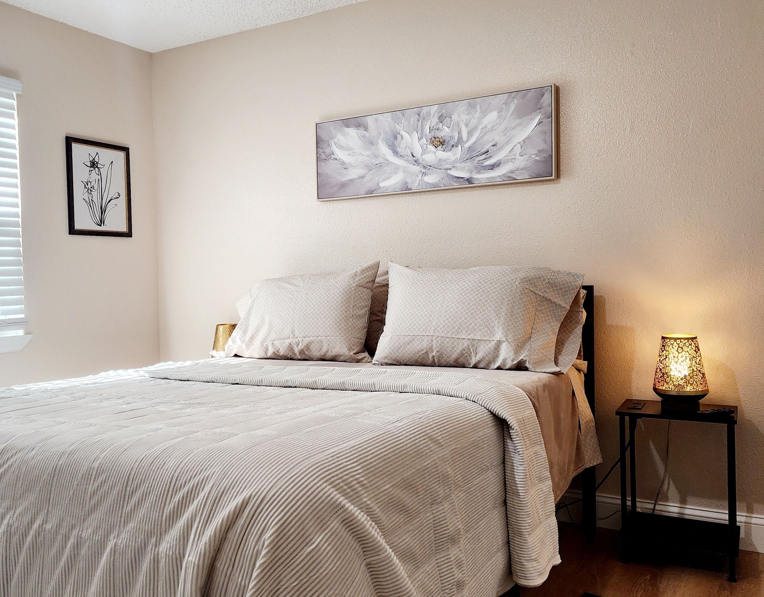 Une chambre à coucher avec un lit, deux oreillers, un tableau floral encadré, une lampe de chevet et une petite table.