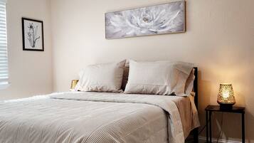Une chambre à coucher avec un lit, deux oreillers, un tableau floral encadré, une lampe de chevet et une petite table.