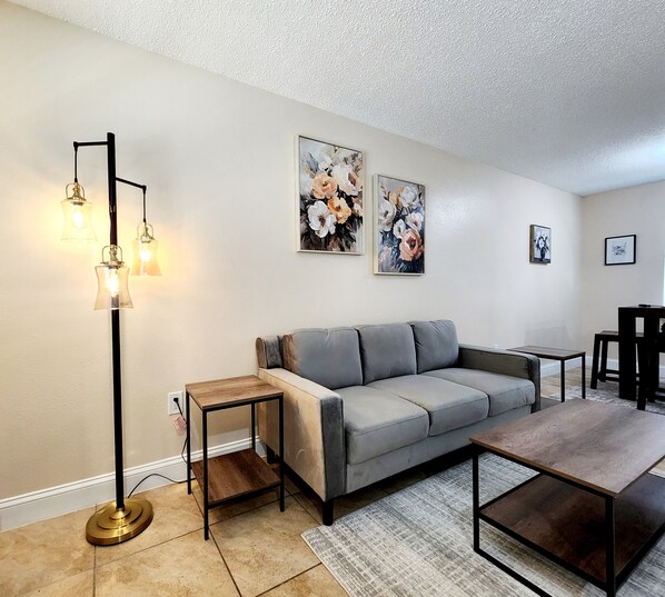 A living room with a grey sofa, a coffee table, a floor lamp, and two floral paintings on the wall. - Toria Suites 922 (Lakeland)