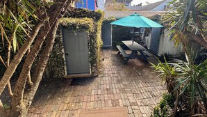 Eine Terrasse mit Holztisch und Bänken, umgeben von üppiger Vegetation und einem blauen Sonnenschirm.