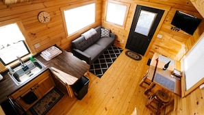 Une petite cabane en bois comprenant un coin cuisine, un canapé, une table à manger et un téléviseur fixé au mur.