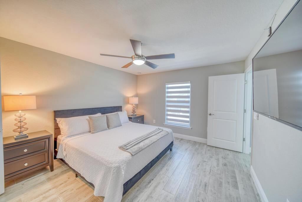 Quarto com cama, mesas de cabeceira, ventoinha no teto, janela com persianas e porta.