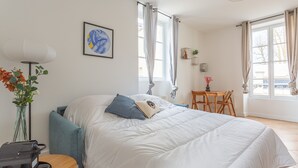 A bedroom with a bed, a desk, a chair, a lamp, a vase with flowers, and a window with curtains.