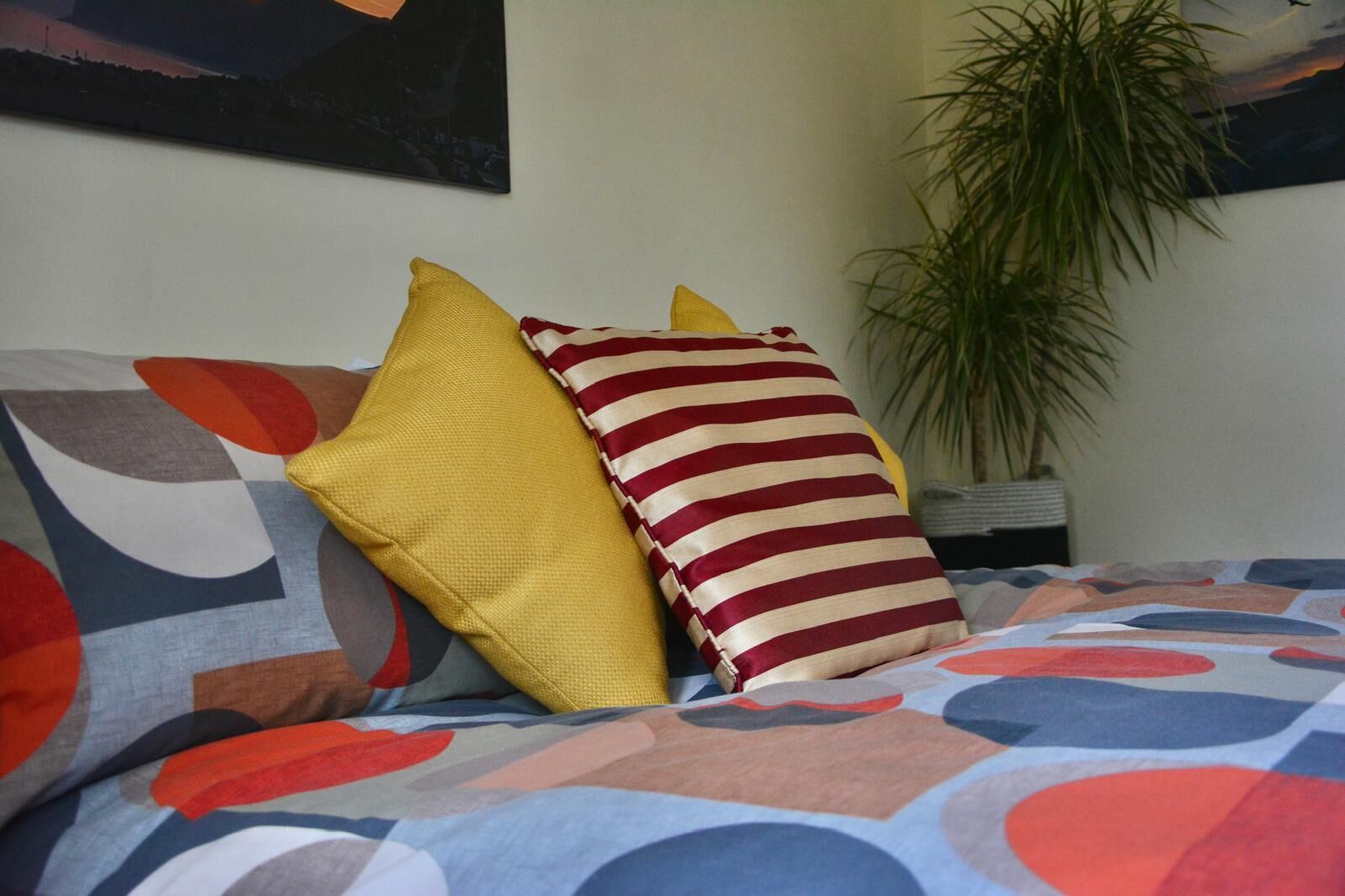 Un lit avec une literie aux couleurs vives et deux oreillers, l’un à rayures et l’autre d’un jaune uni, dans une pièce où se trouvent une plante et un tableau accroché au mur.