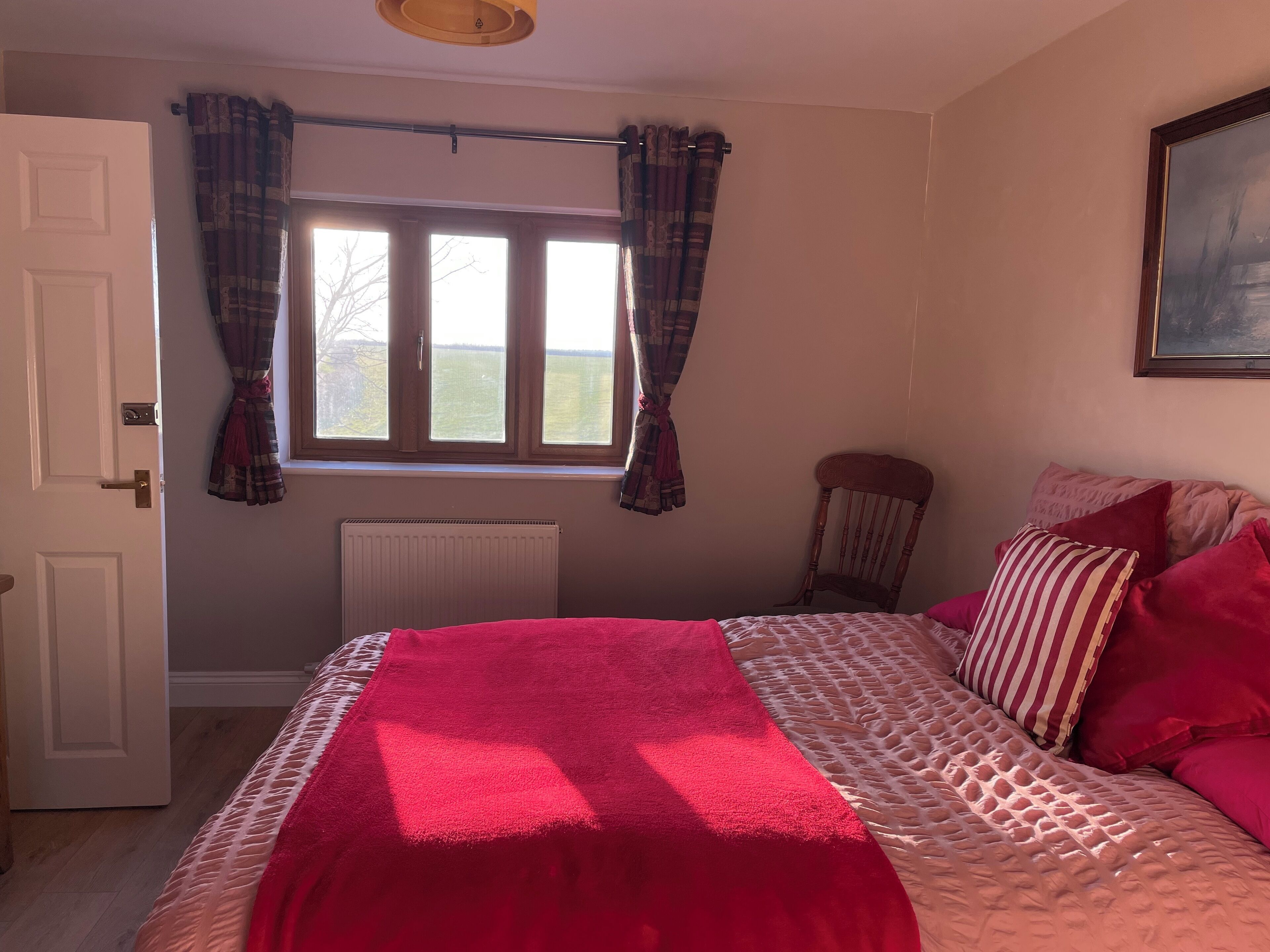 A bedroom with a bed, a radiator, a window with curtains, a chair, and a painting on the wall.
